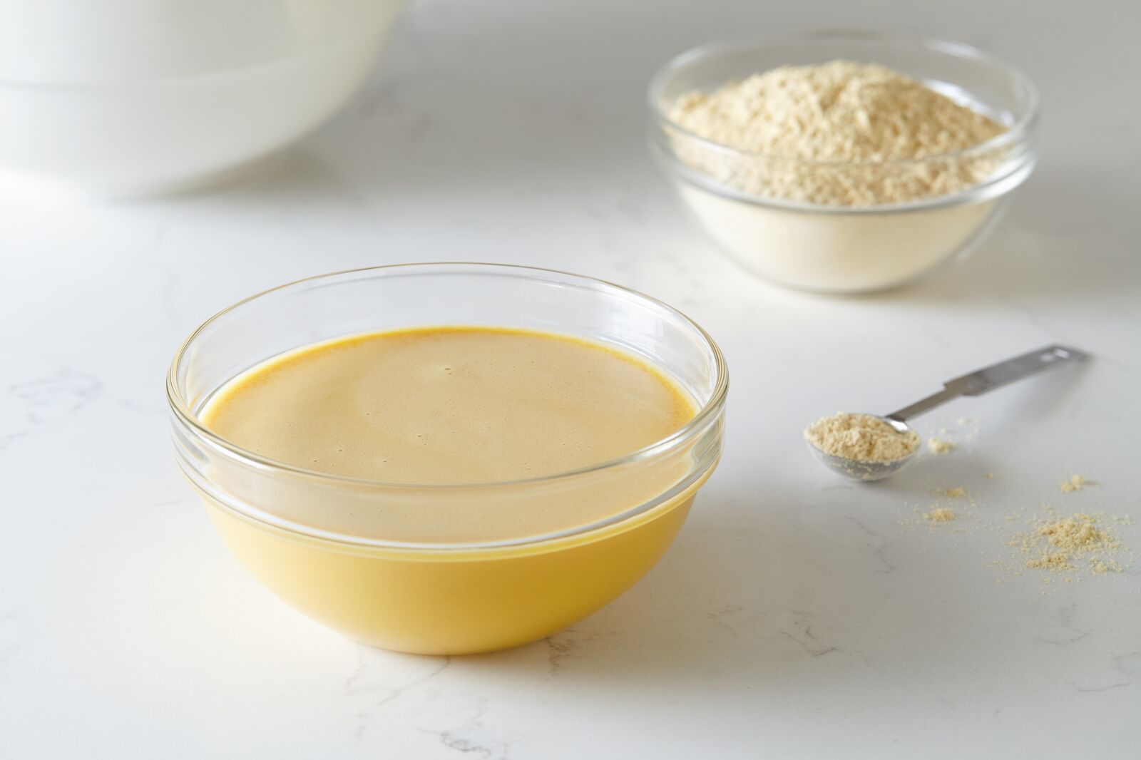 Bowls of egg replace in liquid and powder forms on a table, with a tablespoon of egg replace on the side